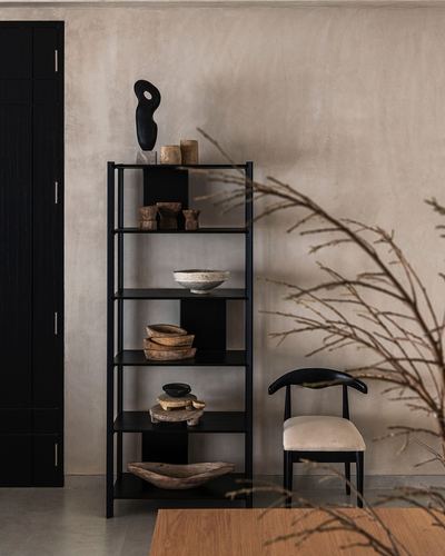 Black metal shelving unit with sculptural objects beside a curved black chair against a plaster wall