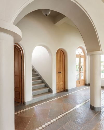 Arched plastered hallway with wooden doors, staircase alcove, and patterned stone floor in Goa home