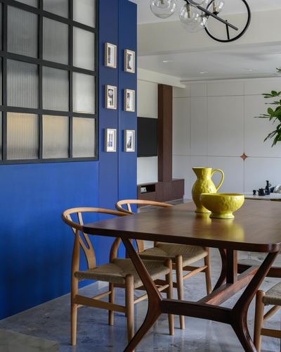 Dining table with wishbone chairs beside a cobalt blue wall and framed art in an open-plan apartment