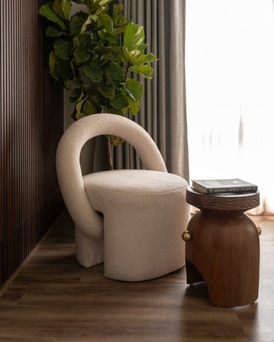 Cream boucle curved-arm chair beside a carved wooden pedestal side table near a window plant