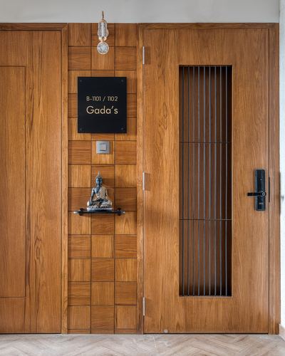 Teak entrance doors with nameplate, small Buddha statue niche, and metal-grille door panel