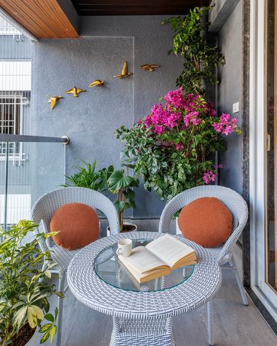 Balcony seating with white wicker chairs, terracotta cushions, glass table, and cascading bougainvillea against grey wall