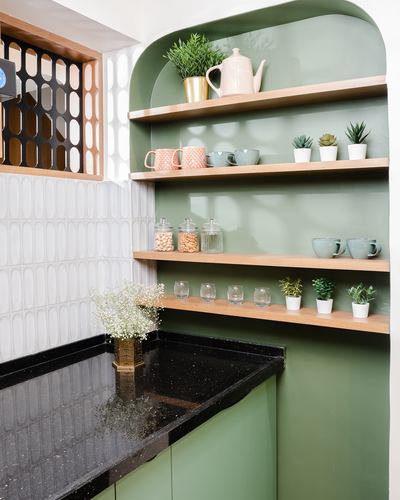 Sage green arched kitchen niche with wooden shelves holding ceramics, plants, and jars above a black countertop