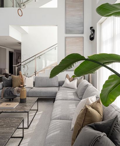 Grey sectional sofa in double-height living room with large plant leaf and glass-railed staircase behind