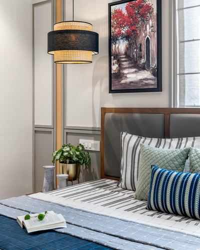 Bedroom corner with wood-framed upholstered headboard, striped bedding, cane pendant light and framed street painting