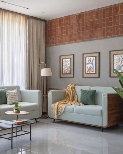 Sage green mid-century sofas below a brick tile and plaster wall with framed folk art