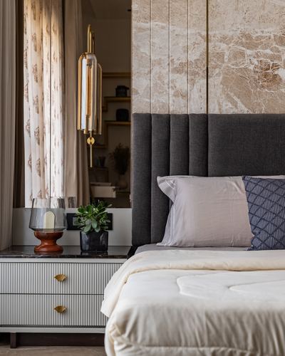 Bedroom with stone-clad headboard wall, charcoal tufted headboard, and brass wall sconce beside mirrored nightstand.