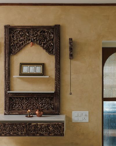 Carved wooden pooja unit with hanging bell on textured ochre wall beside a jaali glass door