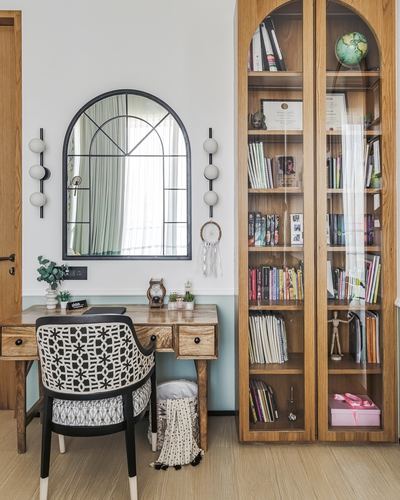 Home study nook with wooden desk, patterned chair, arched mirror, and glass bookcase filled with books