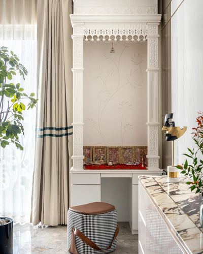 Carved white stone pooja niche with painted panel, brass bell, and houndstooth stool beside marble counter
