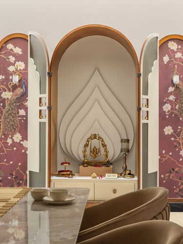 Arched pooja niche with brass idol on swing, flanked by peacock-print wallpaper doors, dining table in foreground