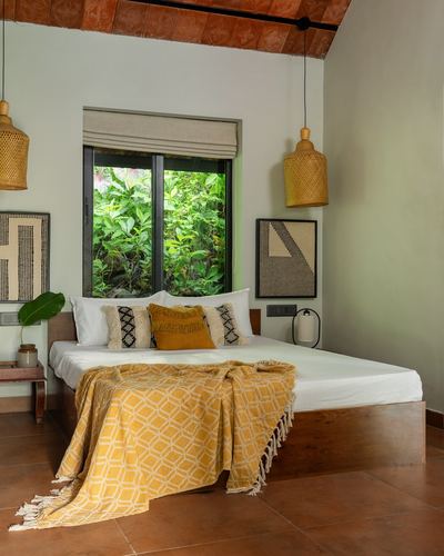 Bedroom with terracotta tile floor, cane pendant lights, wooden bed, mustard throw, window facing greenery