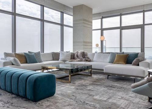 Floor-to-ceiling windows frame a neutral sectional sofa with teal accents and a marble-topped coffee table