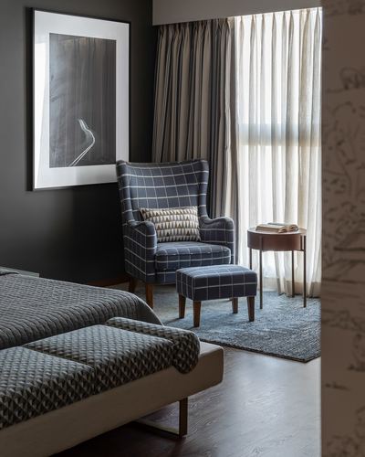 Dark bedroom corner with checked wingback chair, ottoman, and sheer-curtained window
