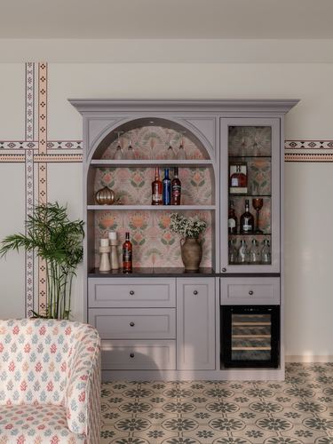 Mauve arched bar cabinet with patterned wallpaper backdrop, glassware, and wine cooler on patterned floor