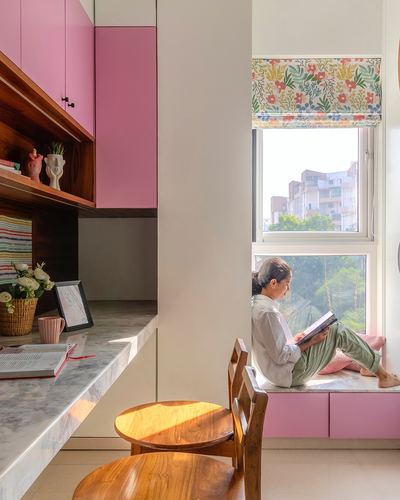 Woman reads on pink window seat beside floral blind in sunlit Pune study nook