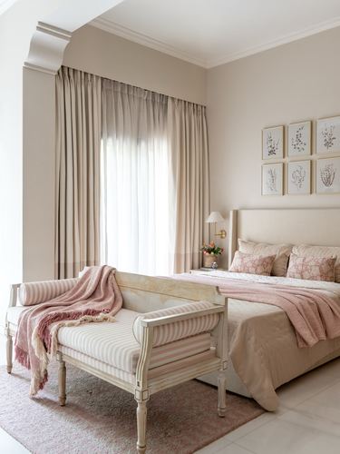 Beige and pink bedroom with upholstered headboard, striped bench, and botanical prints on wall