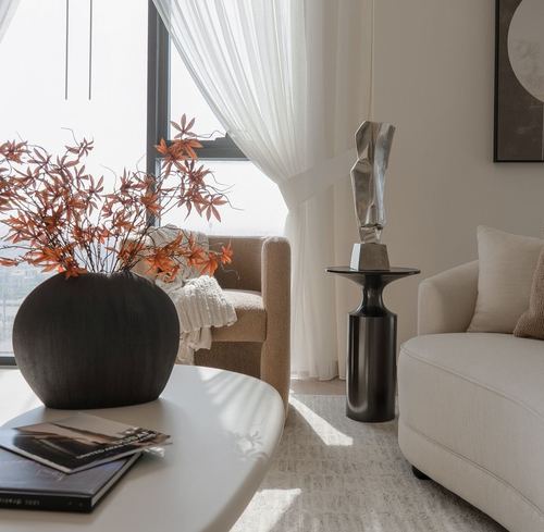 Living room corner with cream sofa, boucle armchair, dark vase with orange branches, and metal side table under sheer curtained window