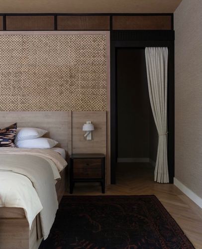 Bedroom with patterned wood headboard panel, cane transom, black-framed curtained doorway, and herringbone floor
