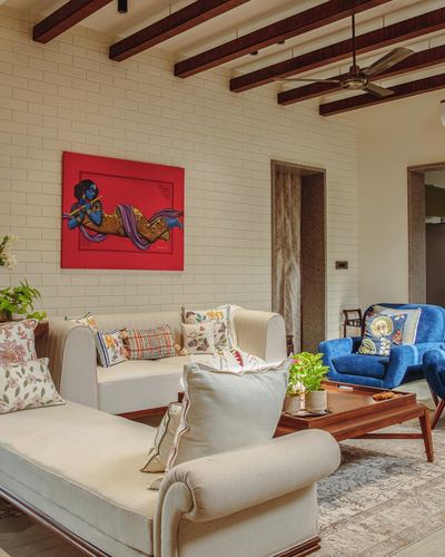Living room with exposed beam ceiling, cream sofas, blue velvet armchair, and red Krishna painting