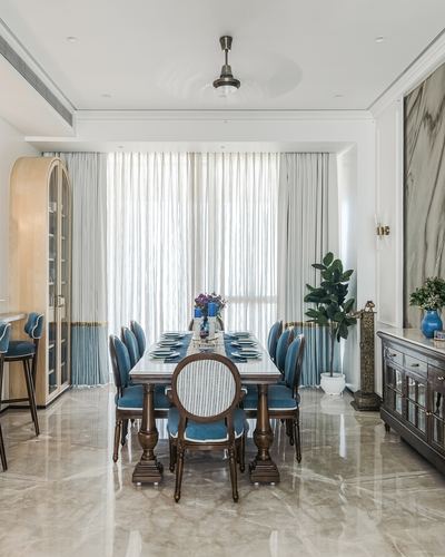 Dining room with blue upholstered chairs, marble table, and arched wooden cabinet near sheer curtained window