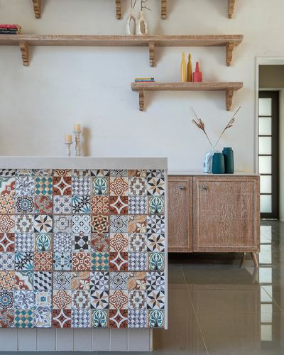 Kitchen island clad in colorful patchwork tiles beneath wooden shelves and a wood sideboard