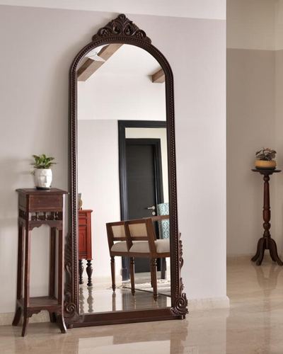 Large carved wooden arched mirror in a foyer, reflecting a cane chair and doorway, beside a wooden console with a plant