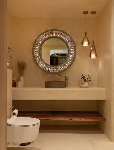 Round textured mirror above concrete sink vanity with wood shelf and amber pendant lights in beige bathroom
