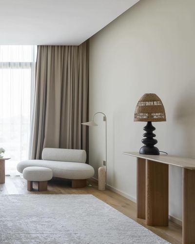 Curved bouclé chaise and ottoman beside sheer-curtained window, wooden console with rattan lamp