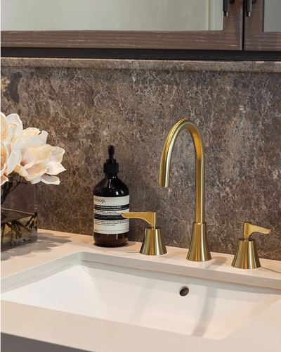 Close-up of gold bathroom faucet and sink against a dark stone backsplash with flowers and hand wash bottle.
