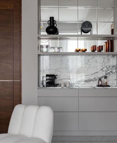 Grey cabinetry niche with marble backsplash and mirrored shelves beside a walnut door and cream chair