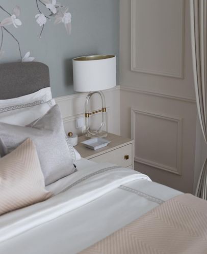 Bedside vignette with white lamp, paneled wall, and layered neutral bedding in soft bedroom