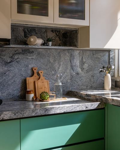 Kitchen corner with grey granite counter and backsplash over teal green cabinetry, styled with chopping boards and vase