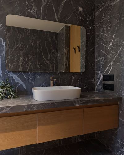 Floating wood vanity with white vessel sink against dark veined stone wall and backlit mirror