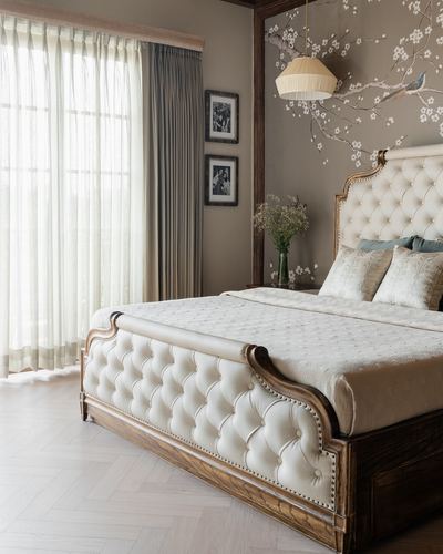 Tufted cream bed with carved wood trim beneath a floral mural and pendant light in a taupe bedroom