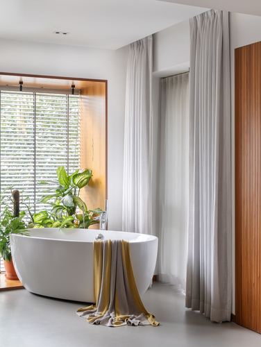 White freestanding tub before a wood-framed window with plants, beside sheer floor-length curtains
