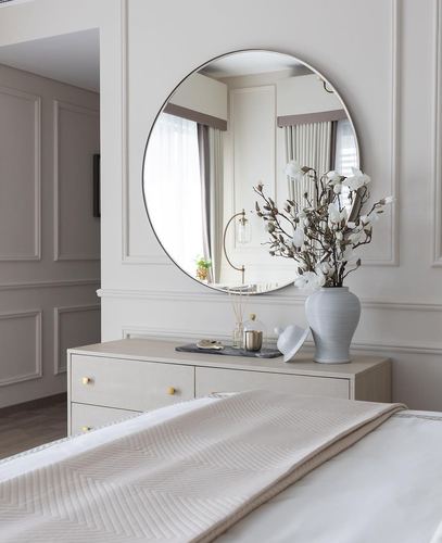Beige bedroom with round mirror above a dresser reflecting a curtained window, vase of white flowers, quilted bed linens.