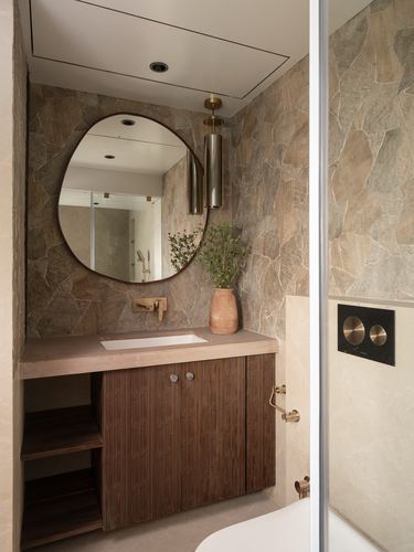 Bathroom vanity with oval mirror, brass pendant, stone walls and fluted wood cabinetry