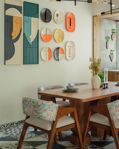Dining table with patterned chairs beneath a wall of decorative face plates and abstract panels