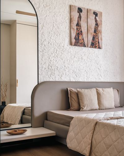 Beige upholstered bed against textured plaster wall with African-motif wall art and arched mirror
