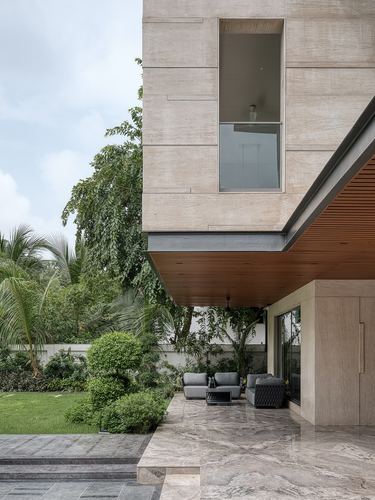 Stone-clad house corner with cantilevered wood-lined overhang above outdoor grey sofas, garden beyond