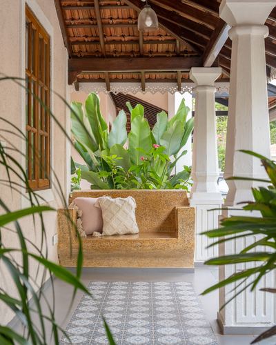 Goan villa porch with a built-in stone bench, patterned tile rug, and lush plants under a tiled roof