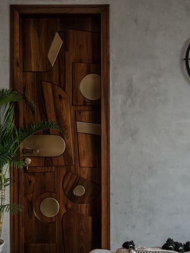 Carved wood door with abstract brass inlay shapes beside a textured plaster wall and palm plant