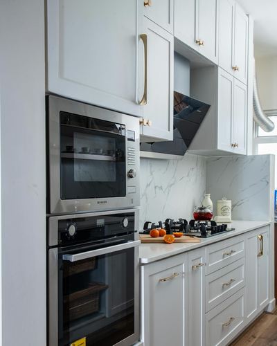 White shaker kitchen with stacked ovens, marble-look backsplash, and gold hardware