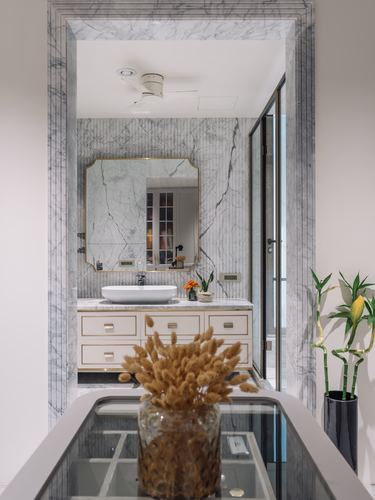 Marble-framed doorway opens onto a bathroom vanity and mirror, seen past a glass table with dried flowers.