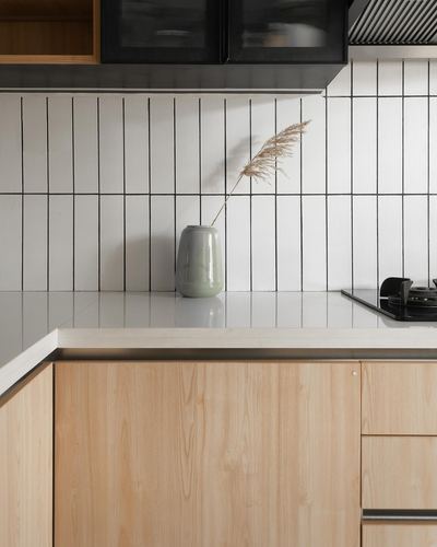 Kitchen corner with fluted white tile backsplash, wood cabinetry, stone counter, and vase with dried grass