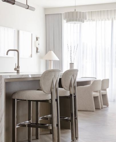 Kitchen island with marble countertop and metal-framed upholstered stools, dining table beyond sheer curtains
