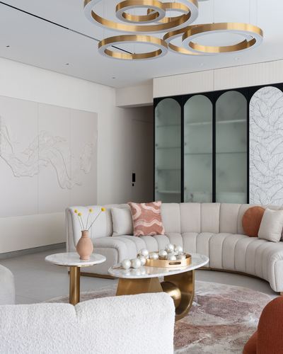 Curved beige boucle sofa beneath a gold ring chandelier, arched glass cabinet behind