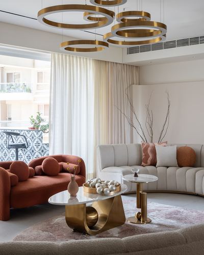 Living room with curved beige sofa, terracotta loveseat, brass and marble coffee tables, and circular gold pendant lights.