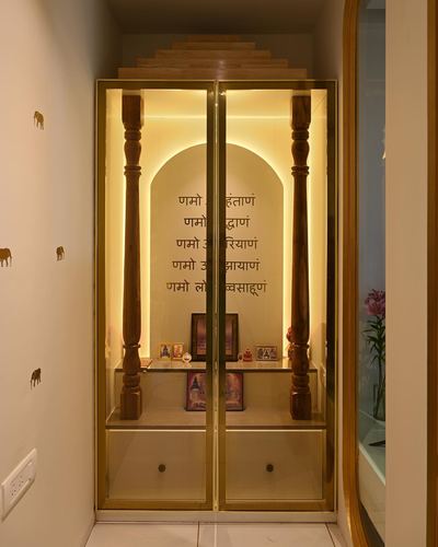 Illuminated pooja unit with arched niche, wooden pillars and brass-framed glass doors in a hallway.